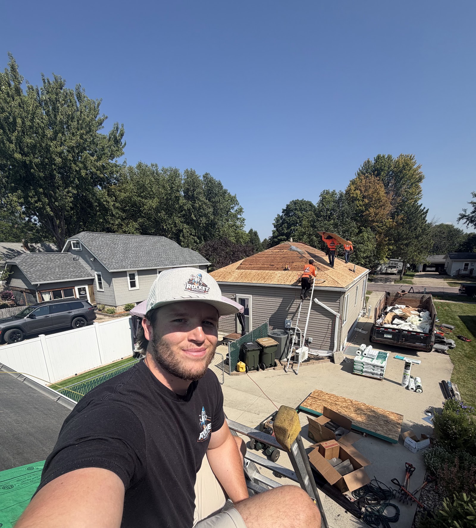Owen on a roof with the crew and dumpster visible in the driveway below