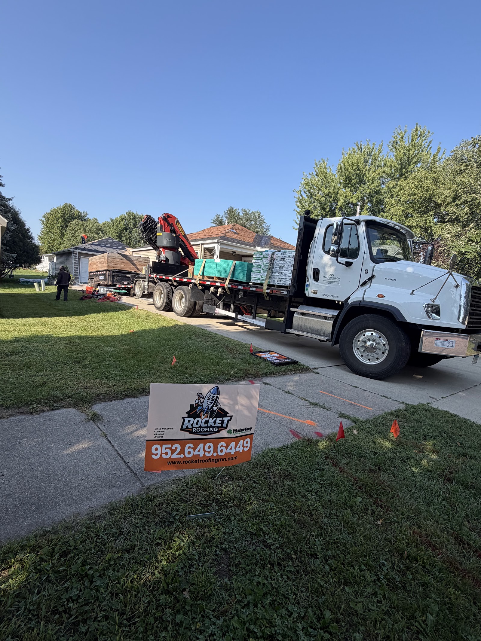 Delivery truck with a boom conveyor unloading shingle bundles, Rocket Roofing yard sign in the foreground