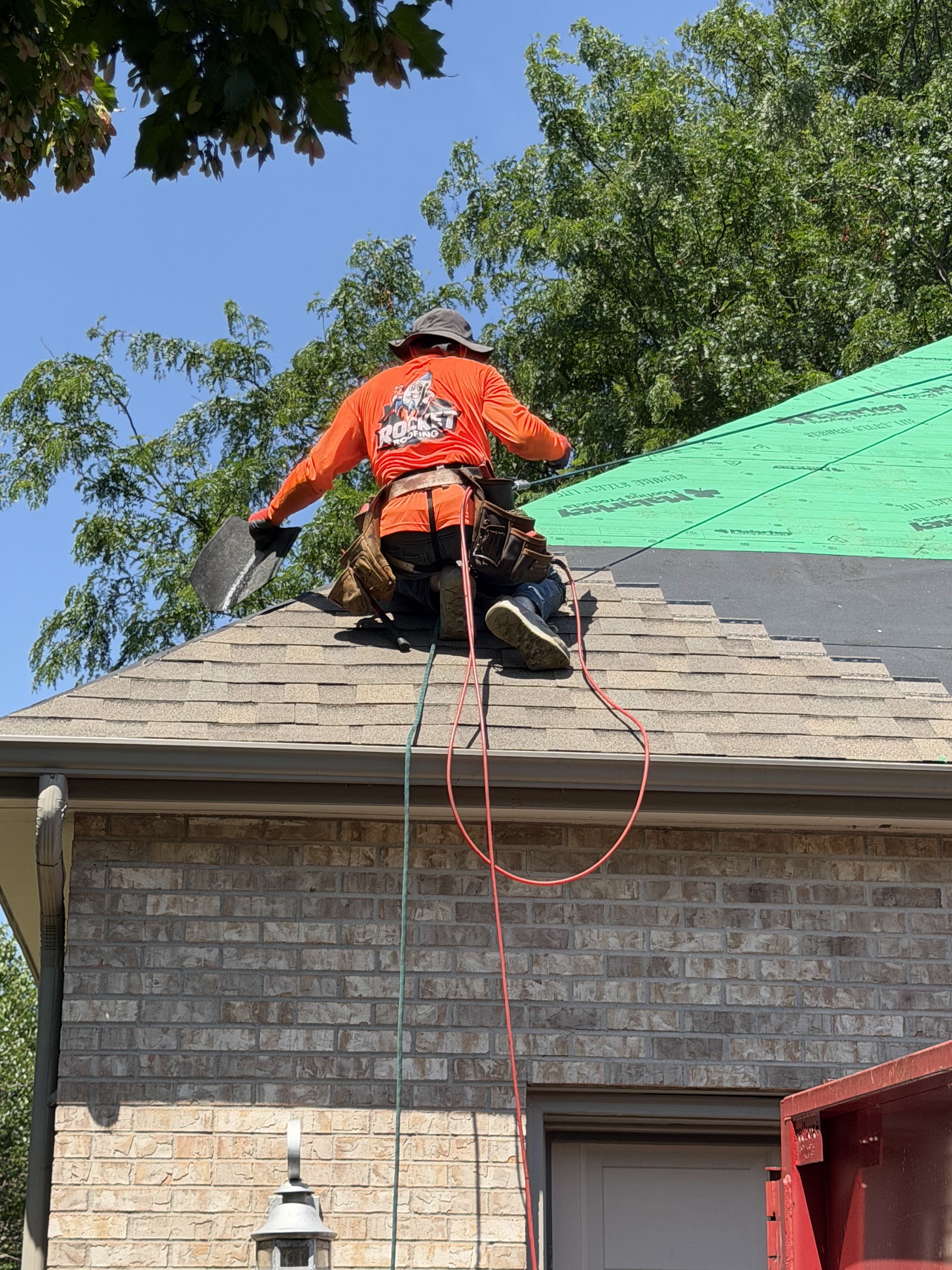 Roofer in a Rocket Roofing shirt harnessed and working the ridge of a tear-off, dumpster below